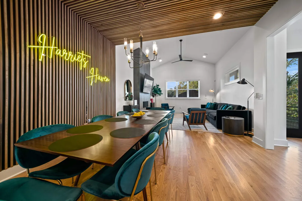This image captures a stylish, open-concept living and dining space with a neon sign reading 'Harriet's Haven' above the dining table. The room features a dramatic chandelier, plush green velvet chairs, and a cozy living area with a comfortable couch in the background. The scene is bright, welcoming, and designed for gatherings.