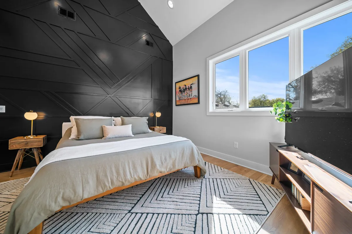 A serene bedroom with a black geometric accent wall, a bed dressed in soft linens, large windows filling the space with natural light, contemporary artwork, and wooden furniture. The overall mood is inviting and calming, offering a tranquil place to relax and recharge.