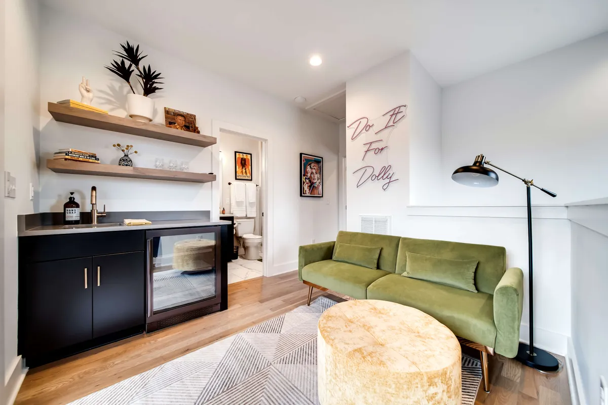 A cozy and stylish indoor lounge setting featuring a green sofa, round coffee table, bar area, decorative shelving, and wall art with text. The space is arranged for socializing and entertaining, with a welcoming mood ideal for intimate gatherings and celebrating special moments in comfort.