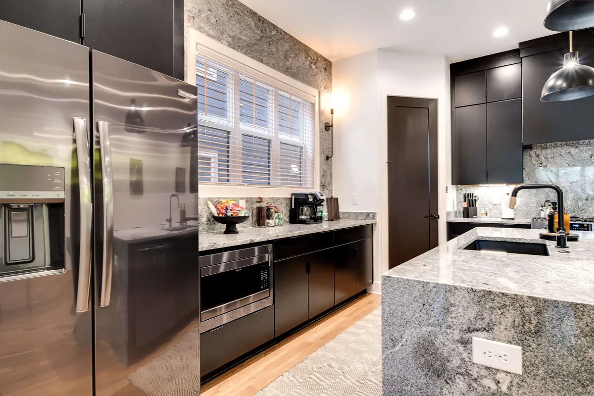 A bright kitchen scene featuring a stainless steel refrigerator, granite countertops, black cabinetry, and a window letting in natural light. The open workspace is perfect for preparing meals and gathering with friends or family.