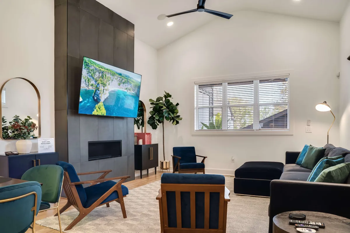 A spacious living room is arranged with modern mid-century furniture, a large television displaying a scenic landscape, and lush green plants. The layout invites relaxation and group conversation, perfect for both family movie nights and entertaining guests.