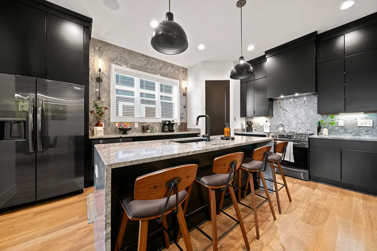 This image showcases a modern kitchen with a spacious island at the center, surrounded by sleek black cabinetry and highlighted by a striking marble backsplash. Stylish pendant lights hang above the island, and the open layout invites both cooking and gathering.
