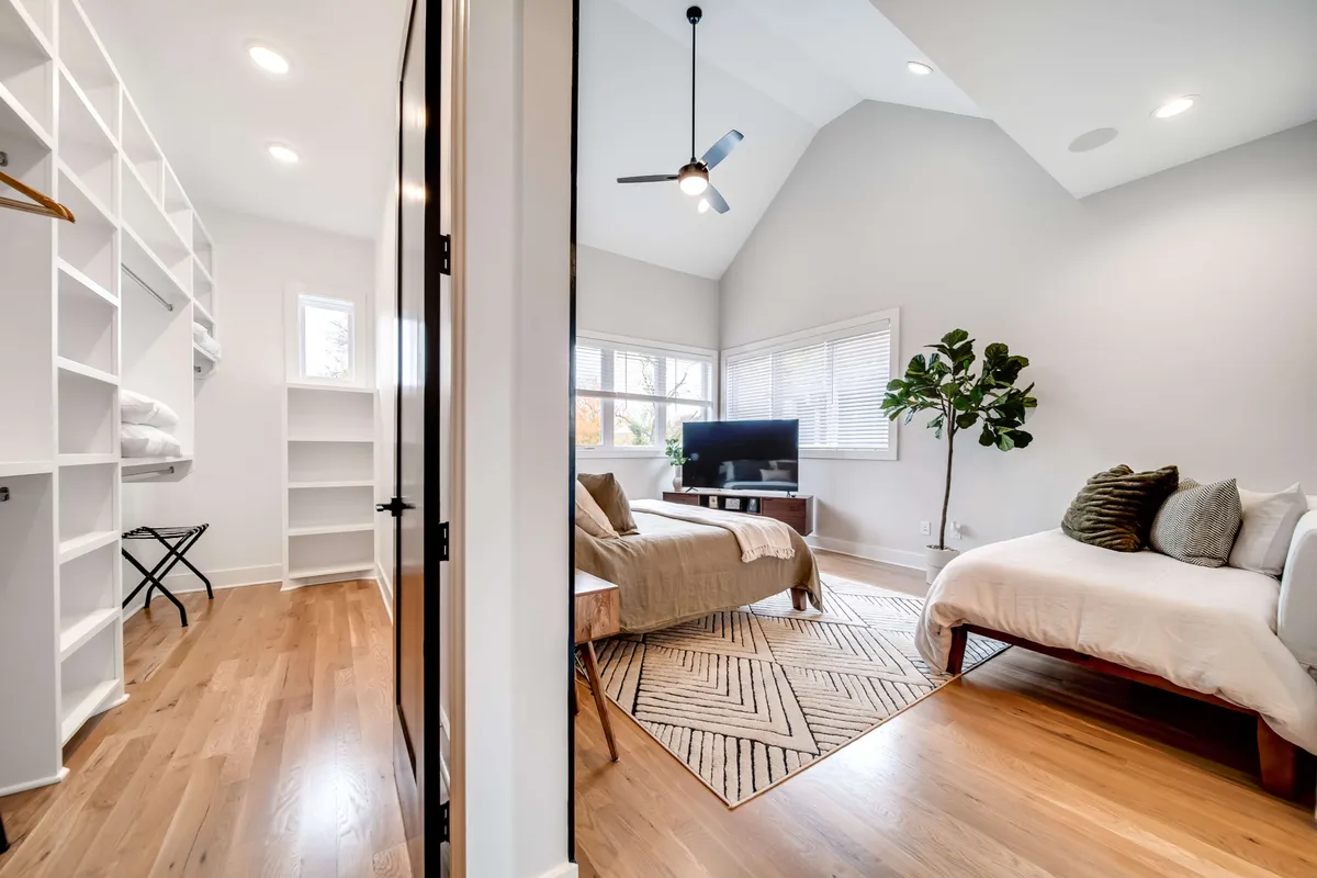 A cozy and stylish bedroom with large windows letting in ample daylight, a neatly made bed with decorative pillows, a minimalist shelving unit, a television on a stand, and a leafy indoor plant adding a touch of nature. The space feels both restful and homely.