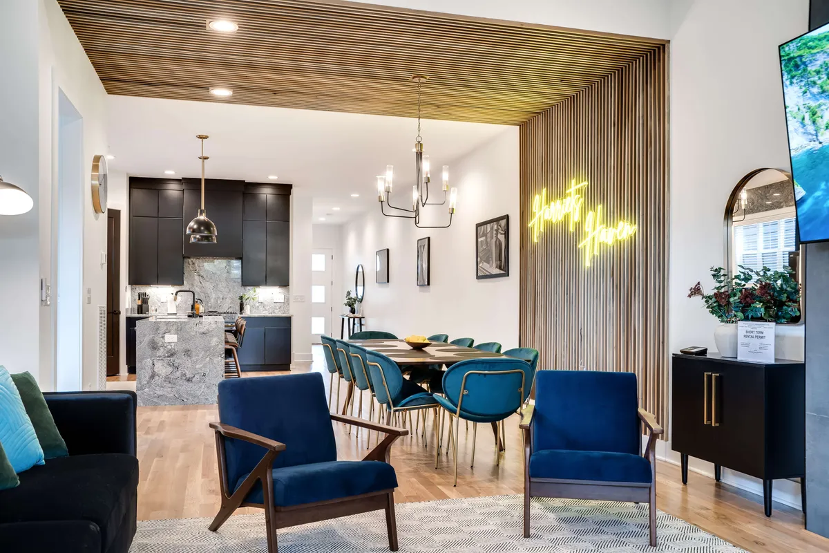A chic dining area with blue chairs around a modern table, illuminated by a playful neon sign and stylish wooden ceiling. The kitchen island in the background hints at an open, communal layout ideal for both intimate and festive gatherings.